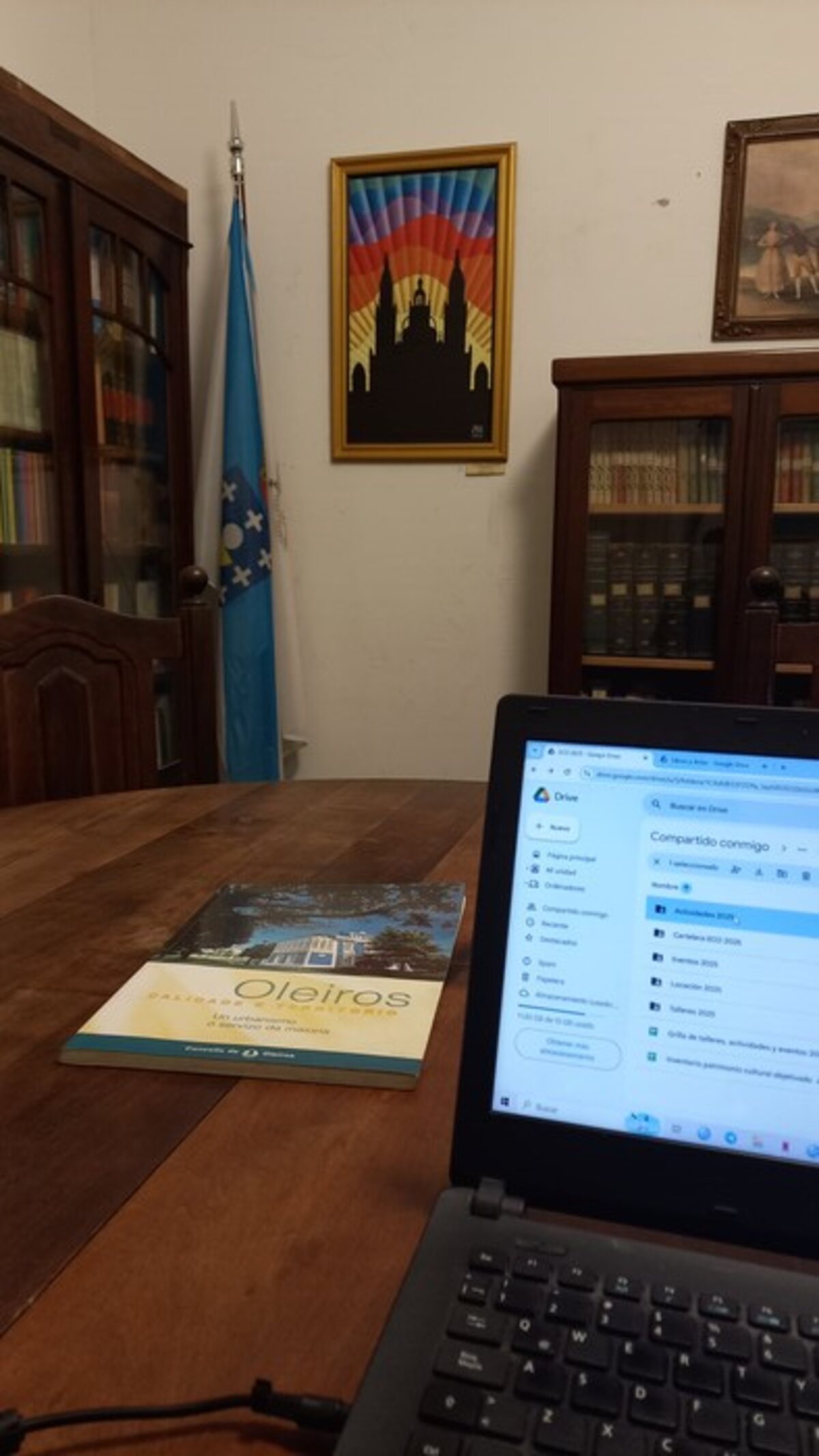 Interior de la sede: bibliotecas de madera con puertas vidriadas, una bandera gallega y un libro sobre el concello de Oleiros apoyado en la mesa.