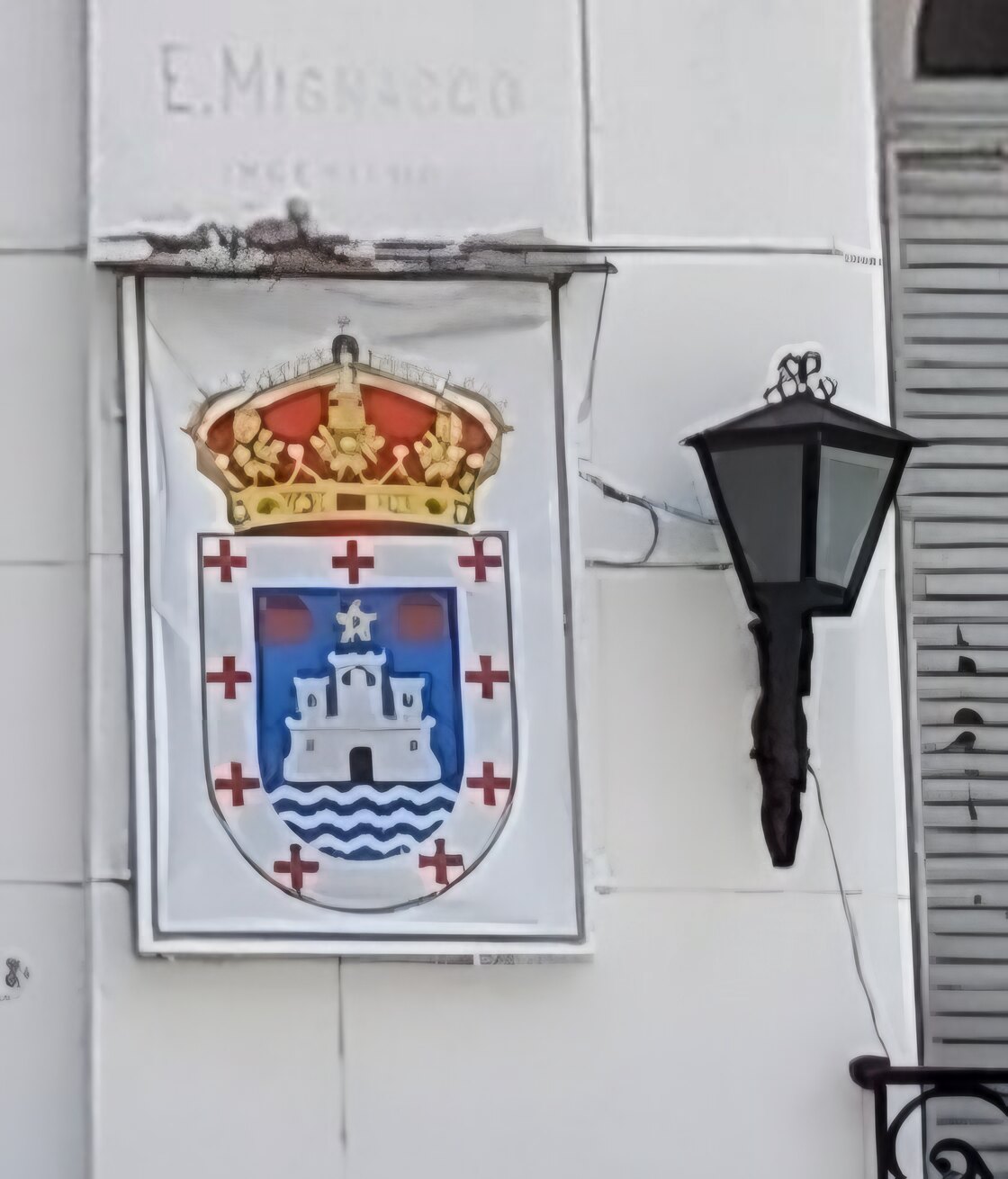 Detalle de la fachada: el azulejo con el escudo de Oleiros (corona real, castillo, olas, estrella y dos ollas de barro) junto a un farol de hierro, bajo la inscripción grabada E. Mignacco, ingeniero.