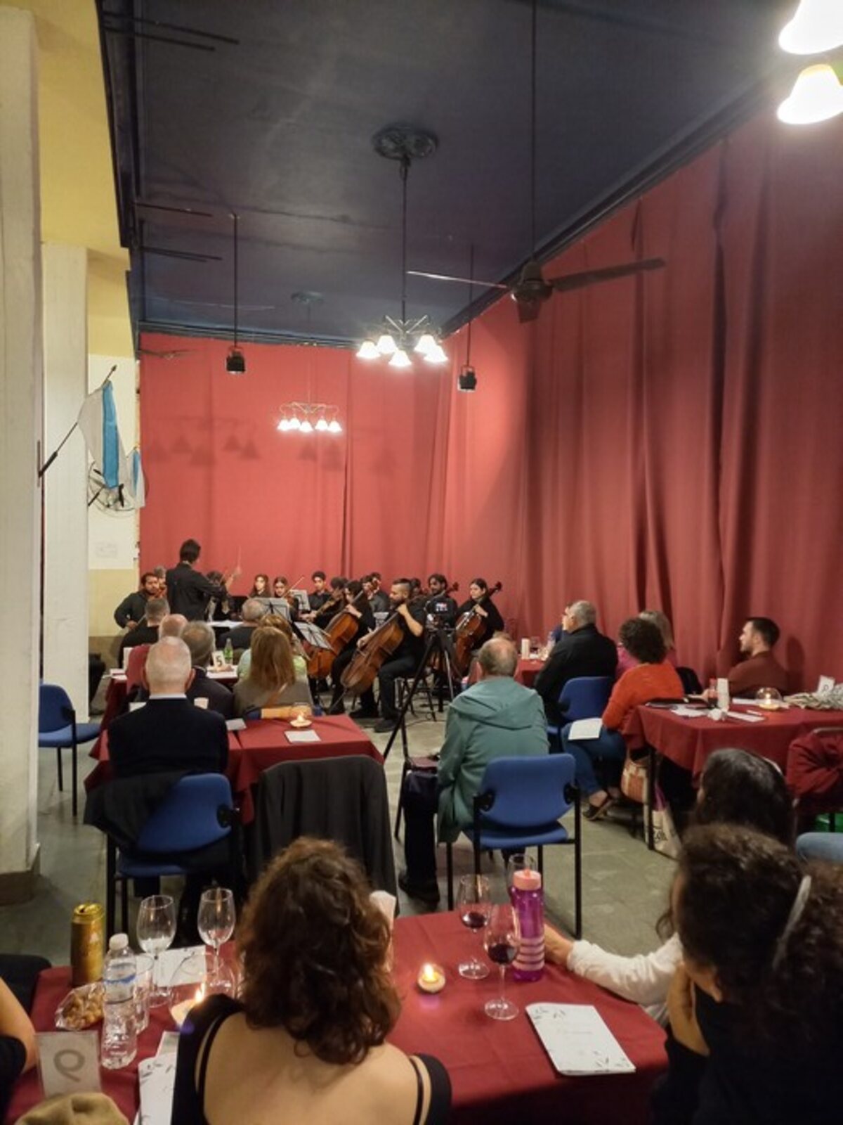El salón durante un concierto: el público sentado a las mesas, de frente a la orquesta, con el telón rojo de fondo.